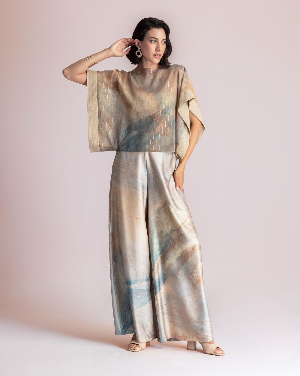 Woman wearing a loose, metallic tie-dye top with wide sleeves and wide-leg satin pants in soft blues, beiges, and peach tones. jewelry visible.