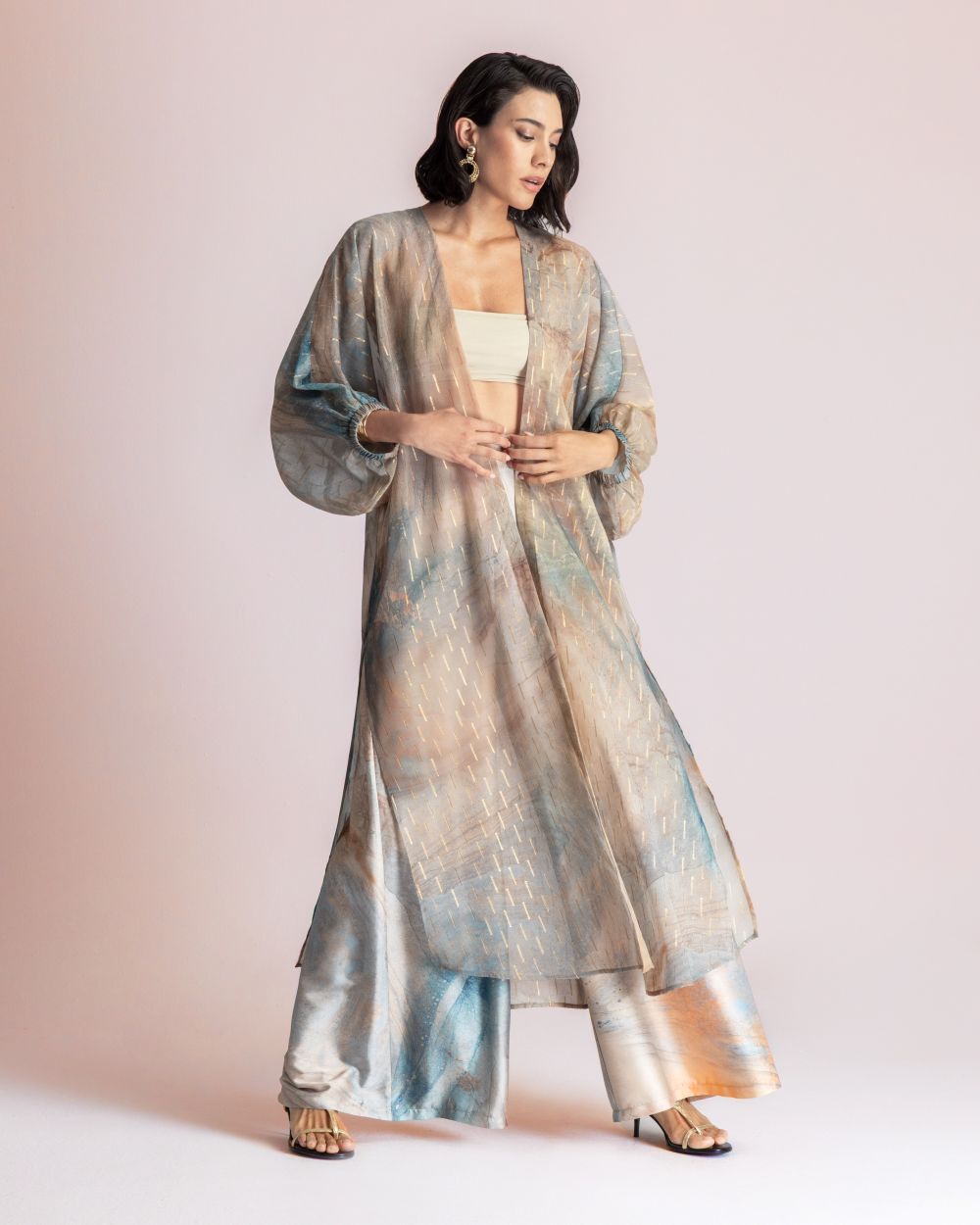 Model wearing a pastel metallic kimono over a beige bandeau and wide-leg satin pants, posed against a pale pink studio backdrop.