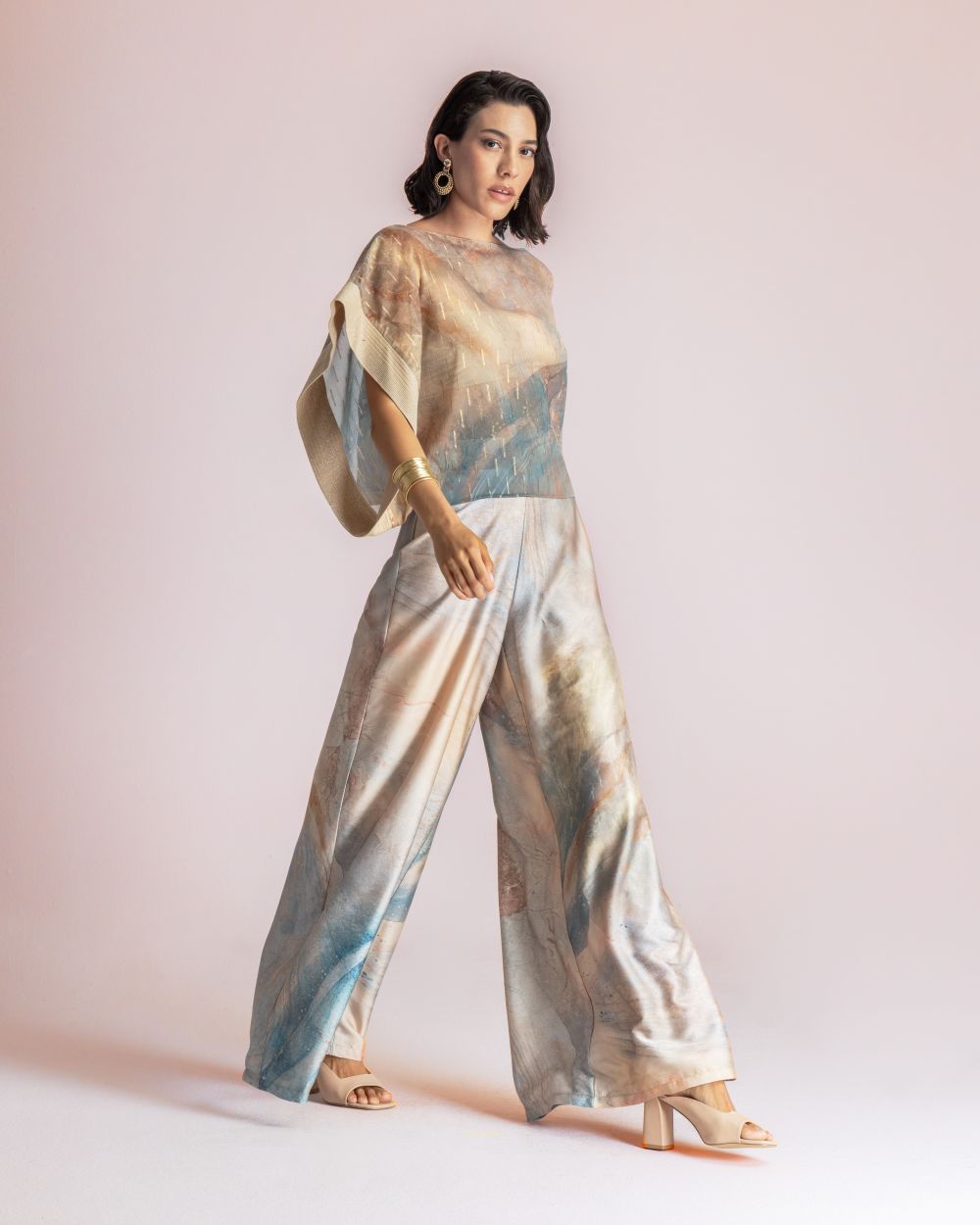 Woman in a pastel tie-dye, wide-leg jumpsuit with flowing cape sleeves, gold earrings, and beige sandals posing against a pale pink background.