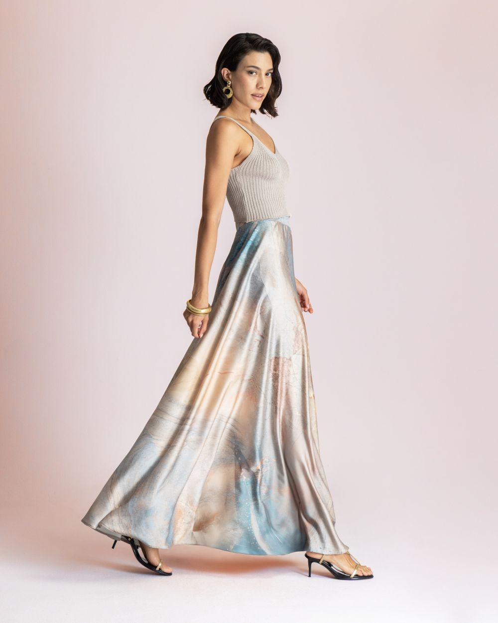 Woman in a light gray knit tank top and a long pastel satin skirt, walking in profile, wearing gold earrings and bracelets.