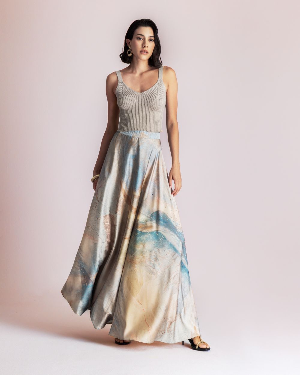 Woman modeling a light grey ribbed tank top with a high-waisted, abstract-paint pastel maxi skirt, standing against a pale pink background.