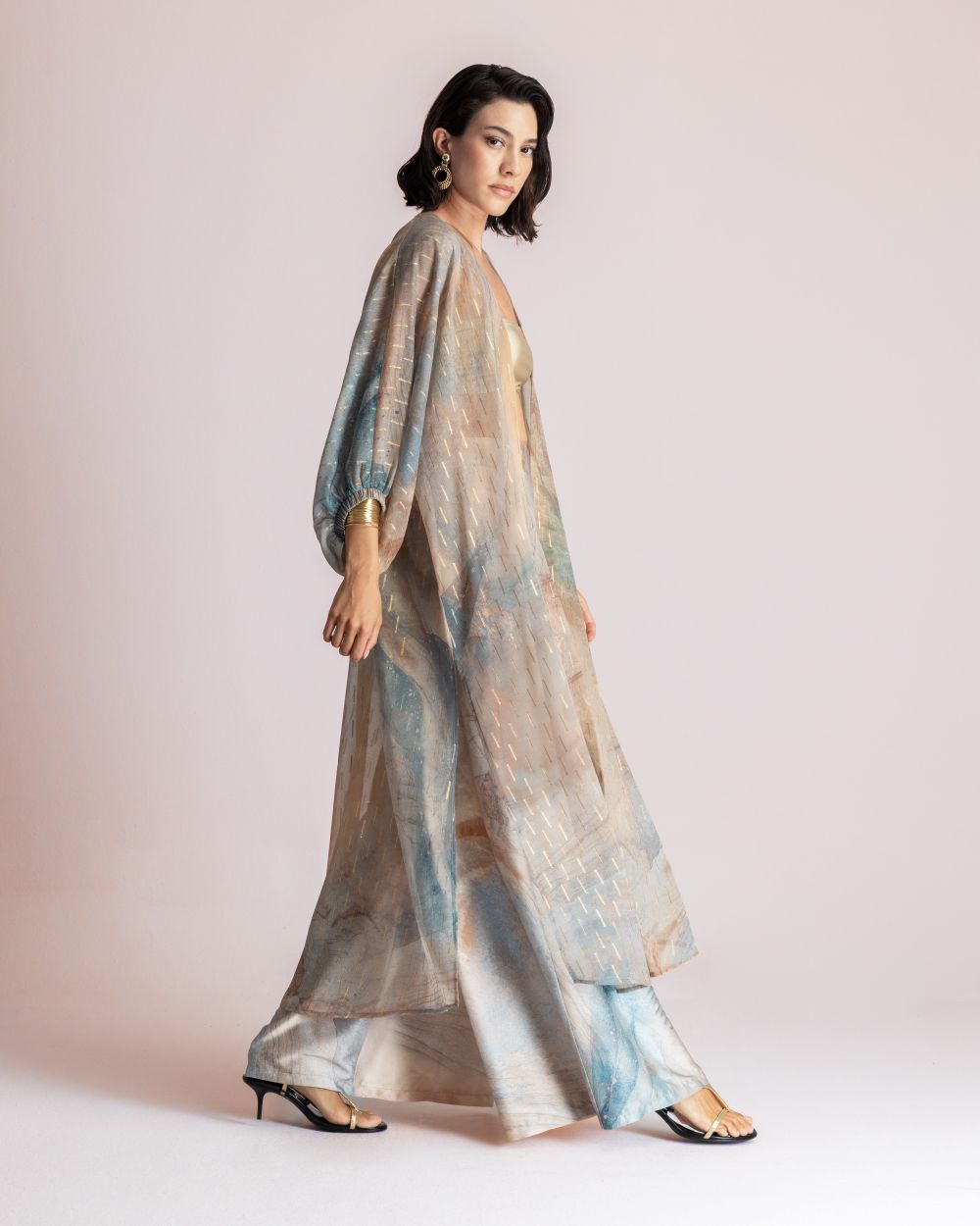 Full-length shot of a woman walking in a pastel tie-dye kurta with matching wide-leg pants, gold earrings, and black sandals, against a light studio backdrop.