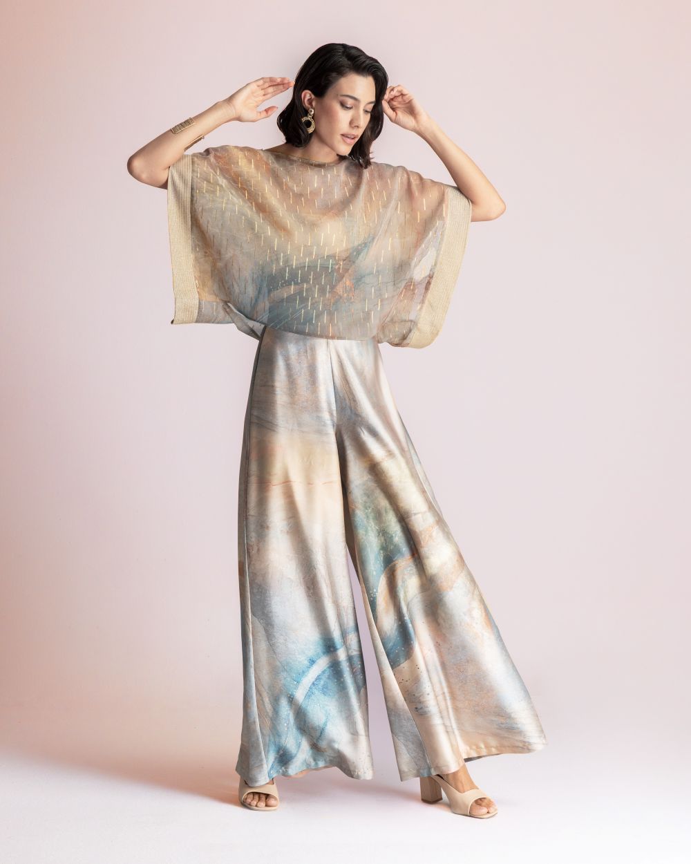 Woman wearing a sheer, beige top with gold stitching over pastel tie-dye wide-leg pants, posing with arms raised against a light pink background.
