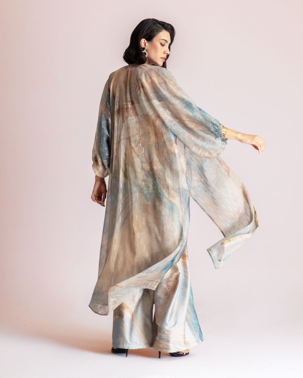 Woman in a loose pastel tie-dye kaftan with batwing sleeves, seen from the back in a studio setting.