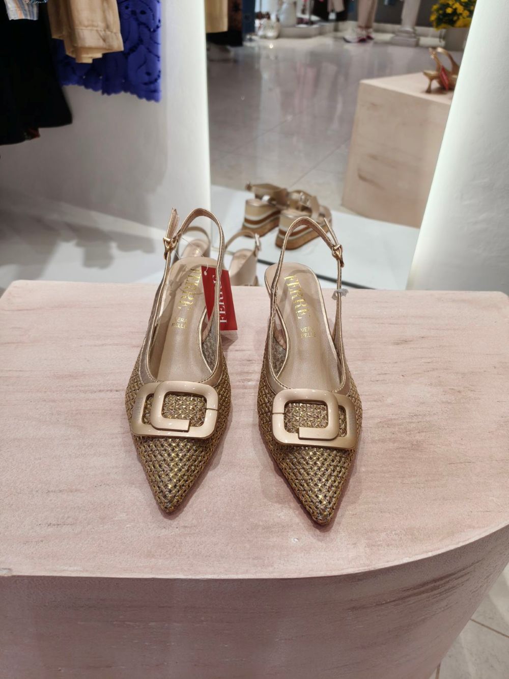 Gold woven slingback heels with a large square buckle displayed on a pink pedestal in a boutique store.