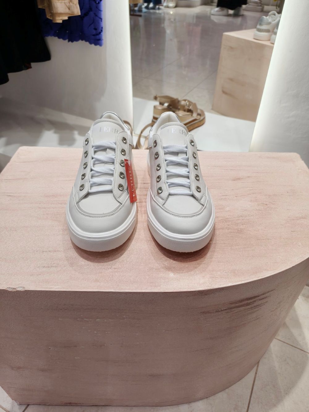 Pair of white sneakers with metal eyelets on a pink display pedestal in a clothing store scenario.