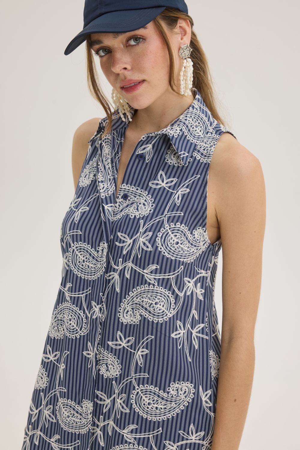 Woman wearing a navy paisley-printed sleeveless dress with a collared neckline and a navy cap, looking at the camera with statement beaded earrings.