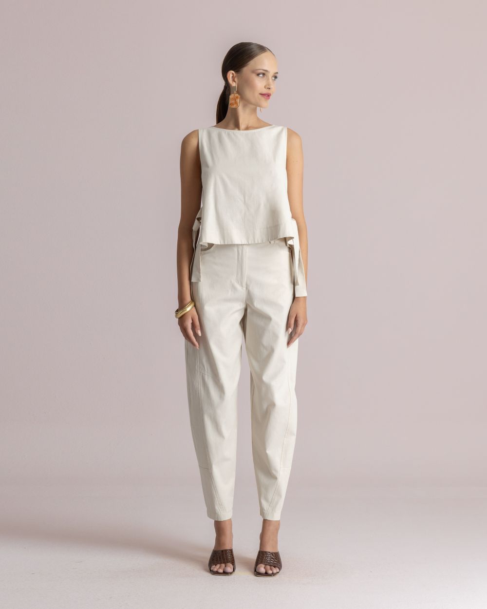Model wearing a sleeveless off-white top and matching high-waisted pants, accessorized with large brown square earrings and gold bangles, standing against a pale pink backdrop.