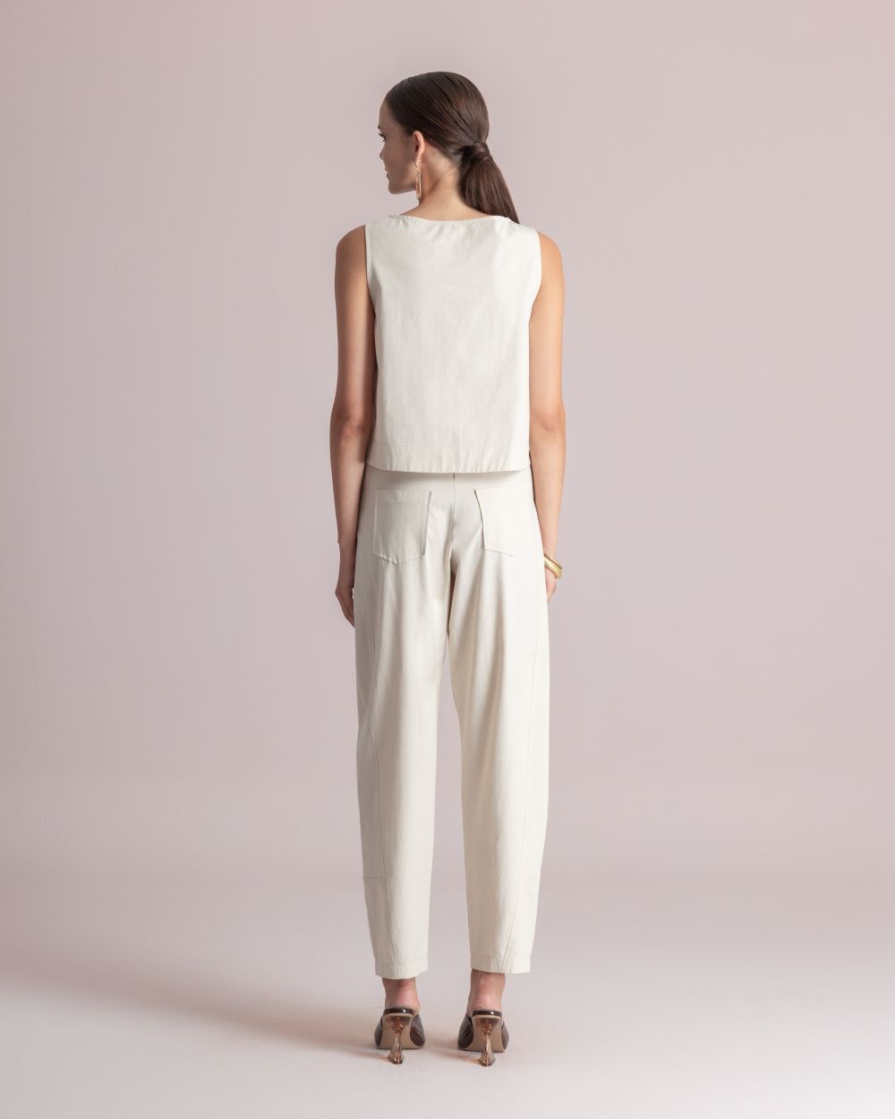 Woman seen from behind wearing a cream sleeveless top and matching high-waisted pants, standing in a pale pink studio with tan heels.