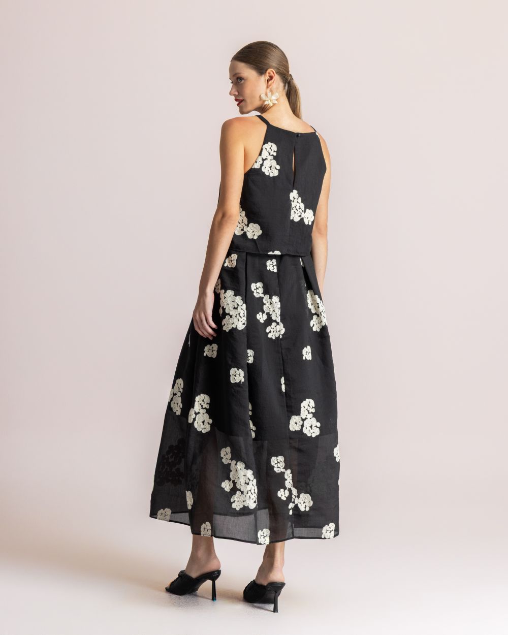 Back view of a woman wearing a black, sleeveless, mid-length dress with white floral embroidery, standing in a studio.