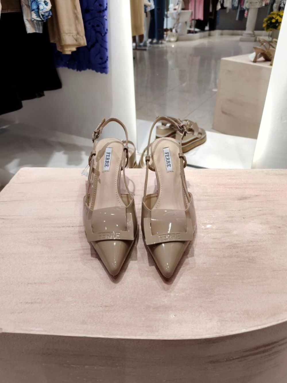 Beige pointed-toe slingback heels with a glossy strap on a display table in a store window; both shoes show 'FERRE' branding on the strap.