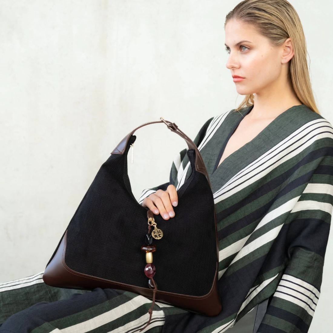 Woman seated in a striped outfit holds a black tote bag with brown leather accents and a beaded charm tassel.