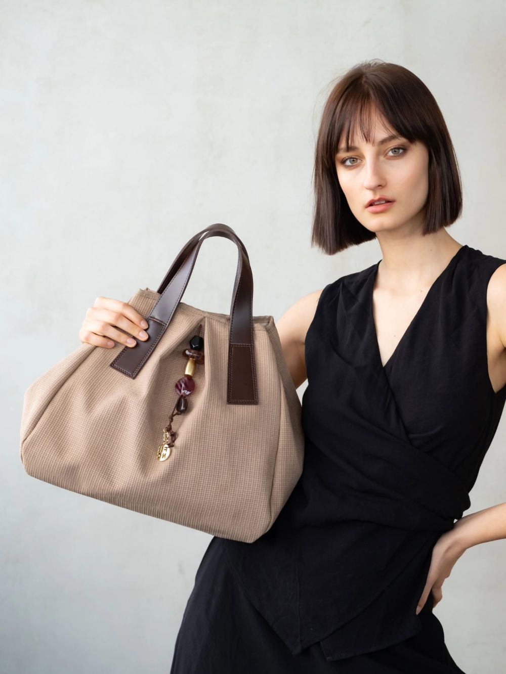 Woman in a black dress holds up a large beige tote with brown leather handles and a beaded charm tag.