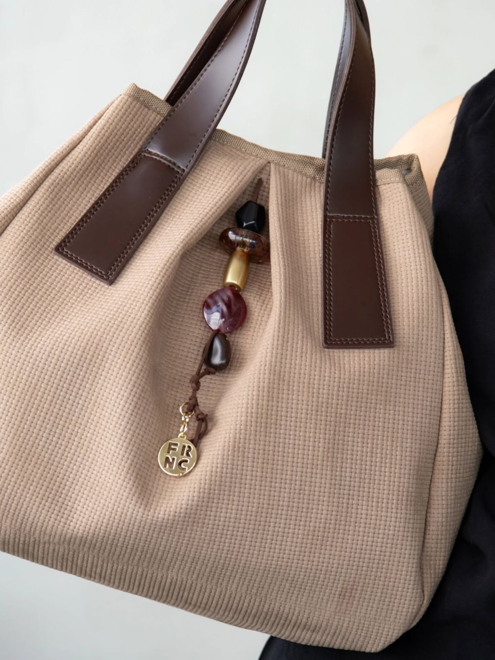 Beige tote bag with dark brown leather handles, featuring a beaded charm and a small gold circular pendant hanging from the strap.