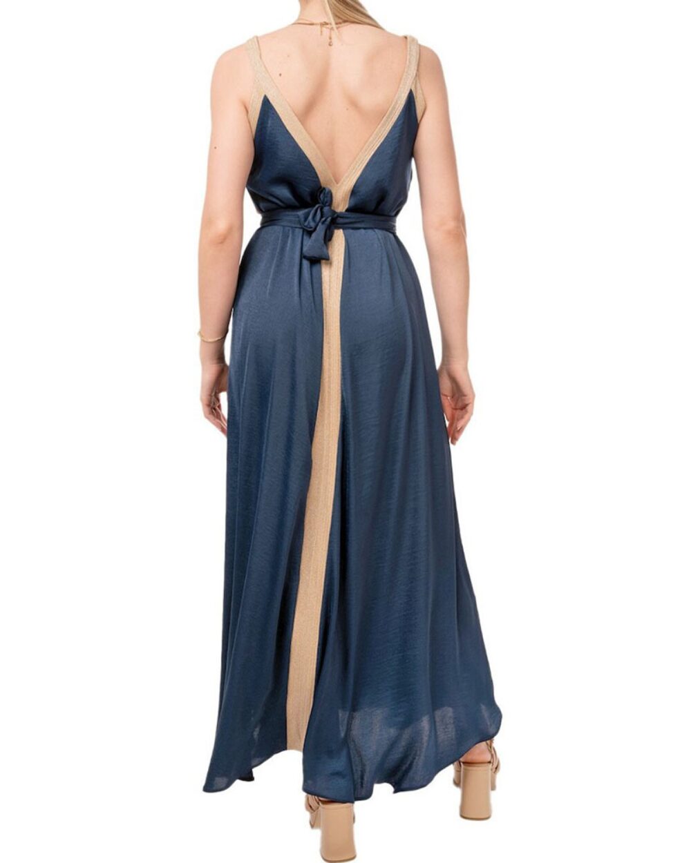 Back view of a woman wearing a navy blue dress with beige trim, open back, and a waist tie, flowing skirt, beige sandals visible.