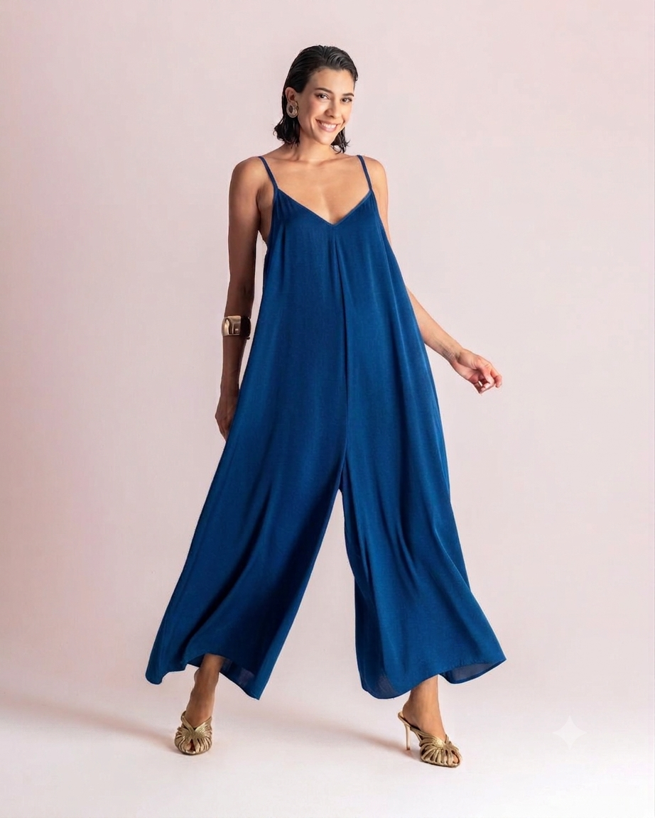 Woman wearing a loose flowing royal blue jumpsuit with thin shoulder straps, posing and smiling against a pale backdrop.