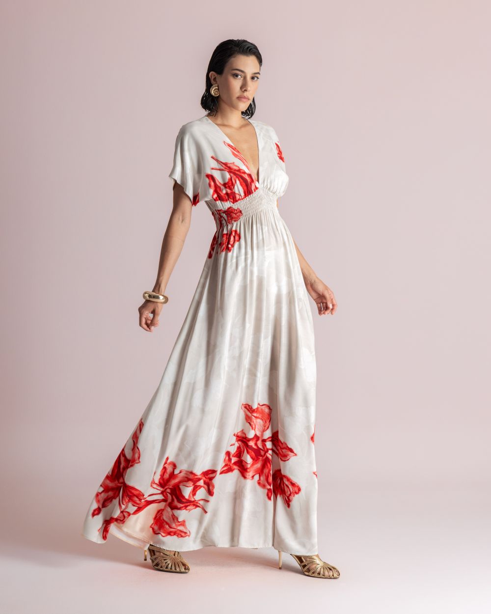 Woman wearing a white maxi dress with red floral print, smocked waist, v-neck, posing against a pale pink backdrop.