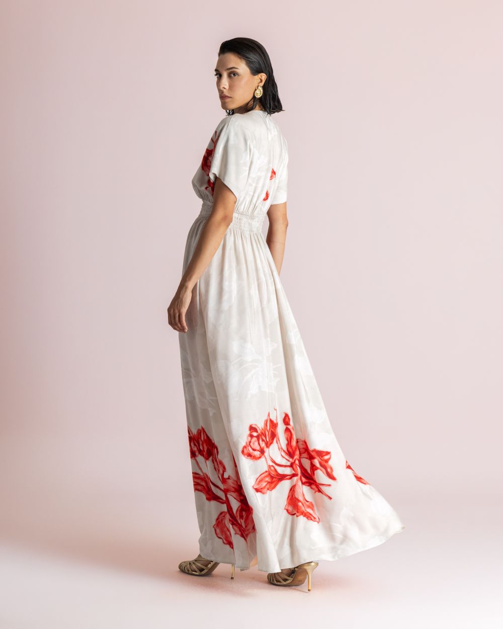 Woman wearing a white maxi dress with red floral print, standing sideways against a pale pink backdrop.