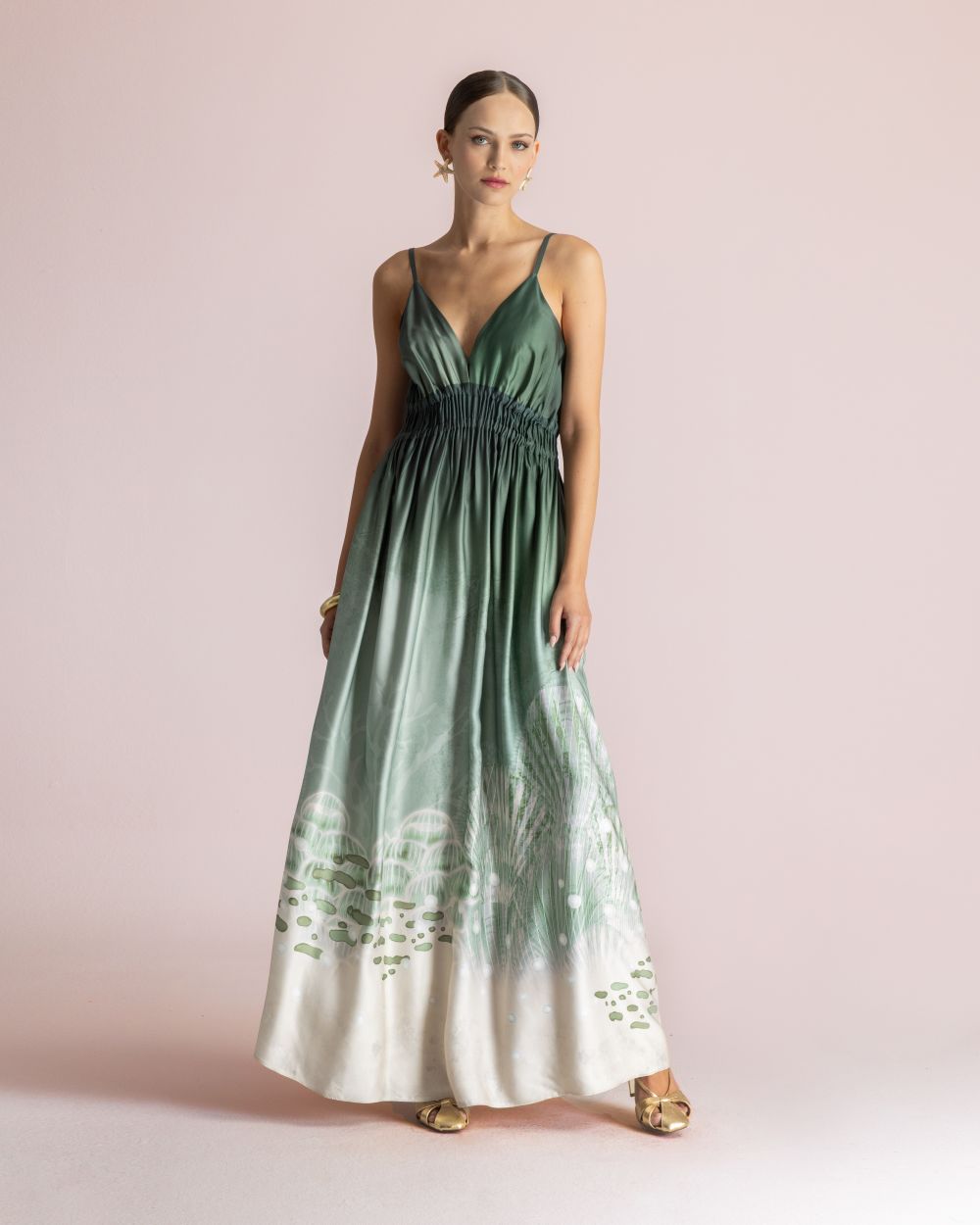 Model wearing a green-to-white ombré spaghetti-strap maxi dress with a cinched, ruched waist and flowing skirt posed against a pale pink backdrop.