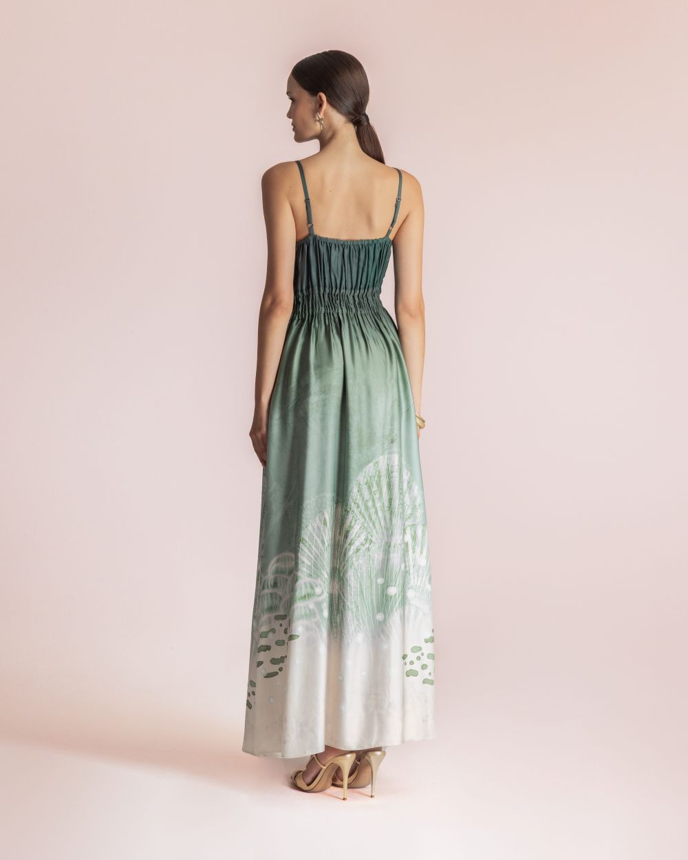 Back view of a woman standing against a pale pink backdrop, wearing a green-to-cream gradient maxi dress with thin spaghetti straps and sandals.