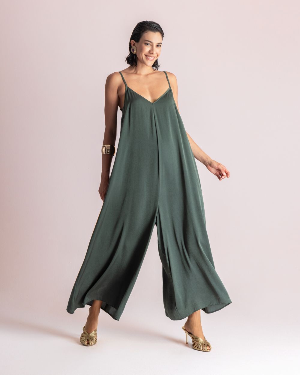 Woman wearing a loose green sleeveless jumpsuit with thin straps, posing against a pale pink background.