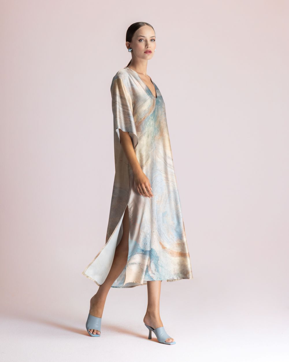Woman modeling a pastel tie-dye maxi dress with a side slit, light blue heeled sandals, and statement earrings against a pale pink backdrop.