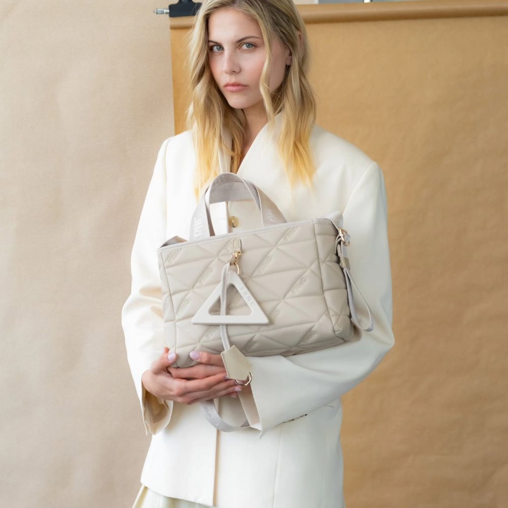 Woman in white blazer holding a light gray quilted handbag with a triangular tag accessory.