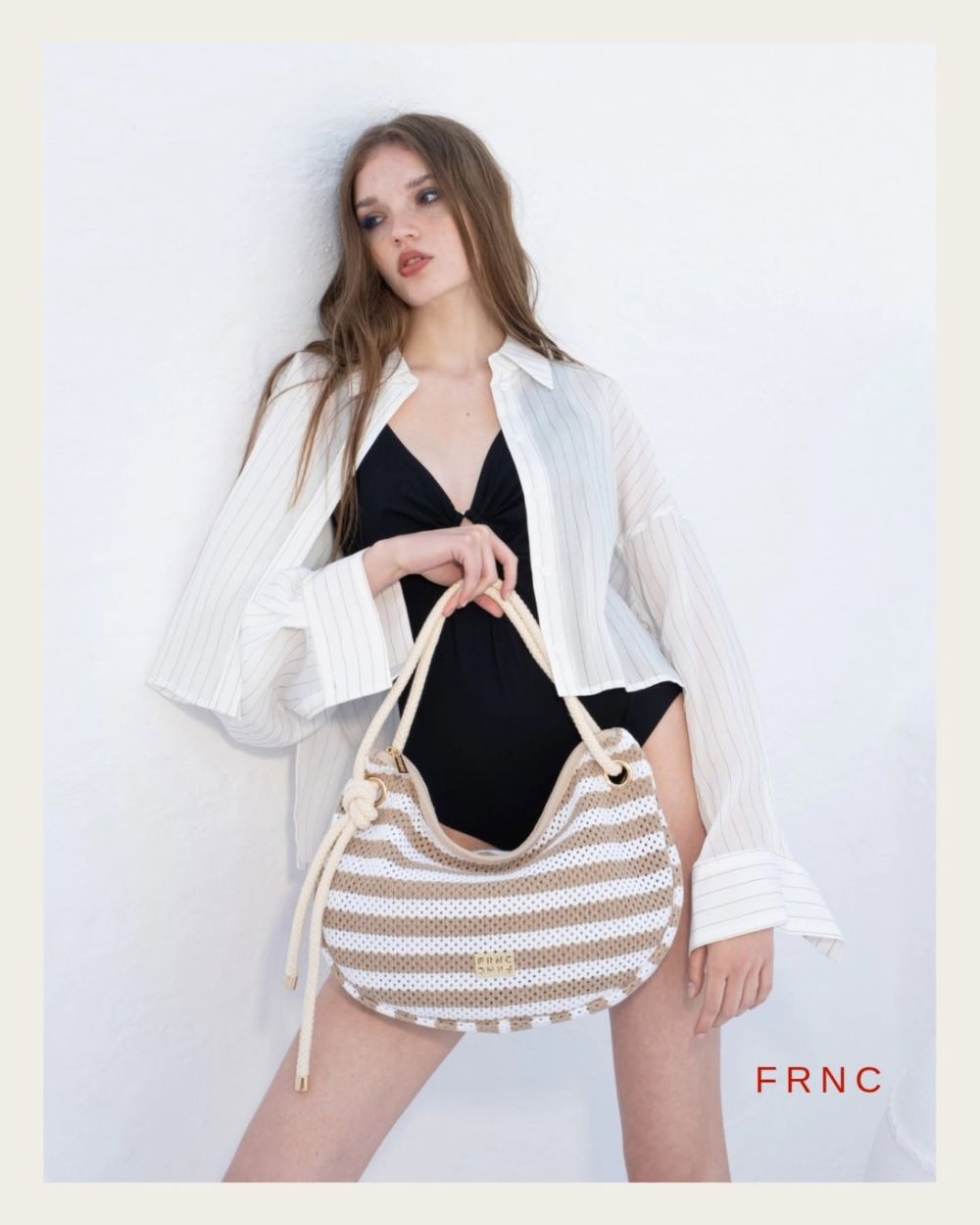 Model wearing a white pinstripe shirt over a black swimsuit, holding a beige striped handbag against a pale background; FRNC logo visible bottom right.