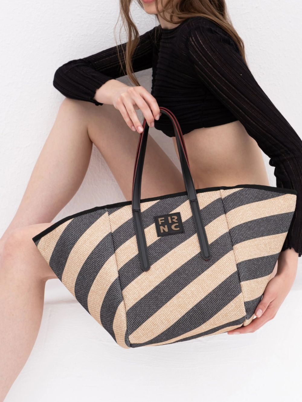 Woman seated against a white wall, holding a large beige and navy striped tote bag with black handles.