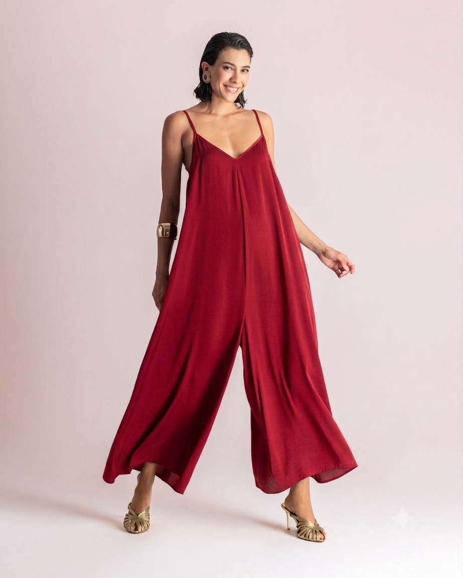 Woman wearing a loose red wide-leg jumpsuit with thin spaghetti straps, posing in gold sandals against a light background.