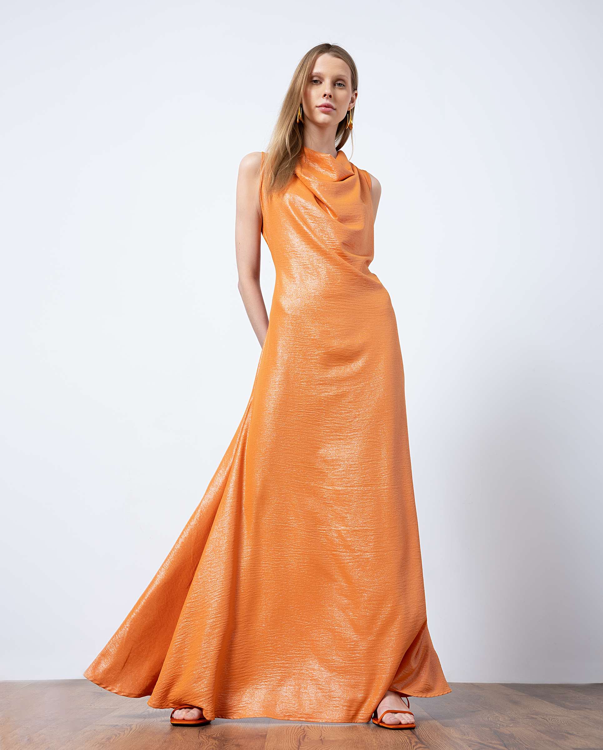 Female model in a shiny orange gown standing, dress sweeping the floor in a studio with a pale backdrop and sandals visible at her feet.
