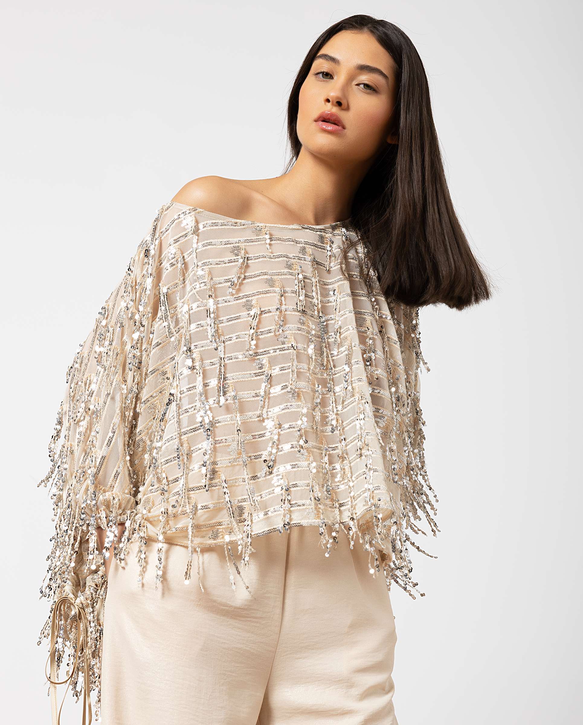 Model wearing a beige off-shoulder sequin fringe top, posing against a light gray background.