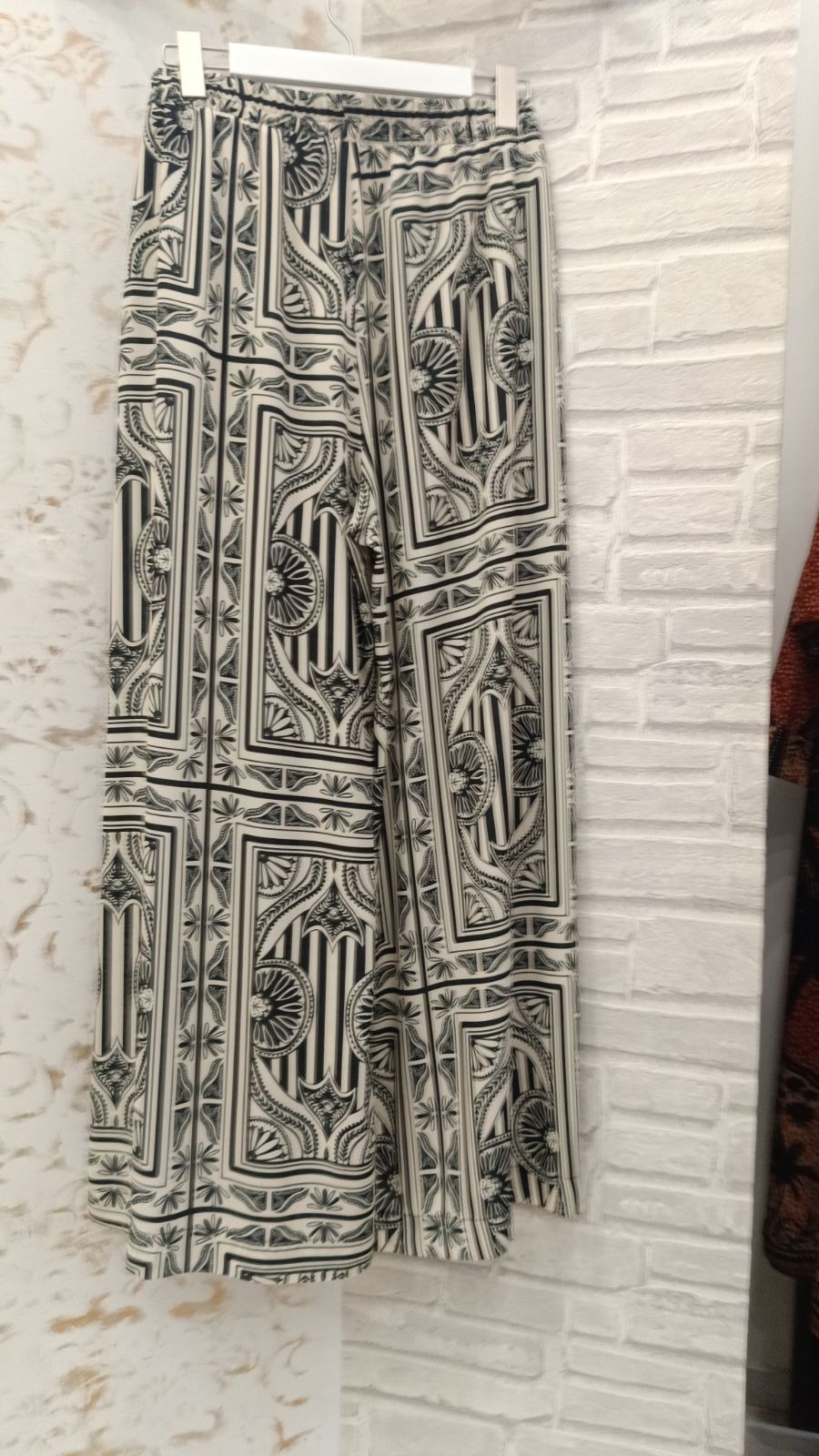Black and white patterned wide-leg pants hanging on a white hanger against a white brick wall.
